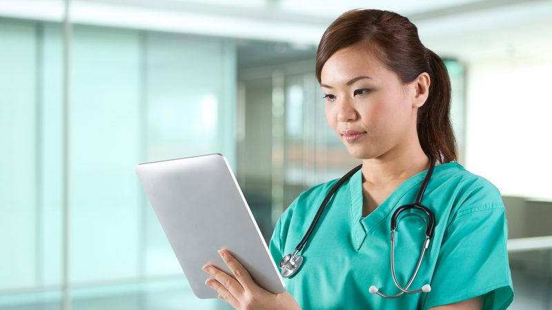 Female Asian doctor looking at a digital tablet & wearing a green scrubs plus stethoscope.